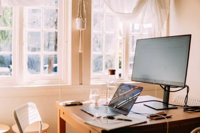 Een zonnige thuiswerkplek met een laptop op een houten bureau en veel natuurlijk licht dat door de grote ramen naar binnen stroomt.